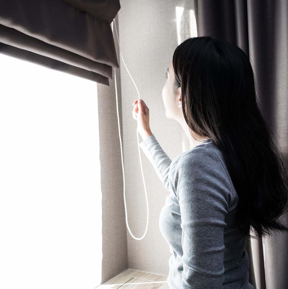 Woman pulling a cord to adjust a gray Roman shade on a sunny window.