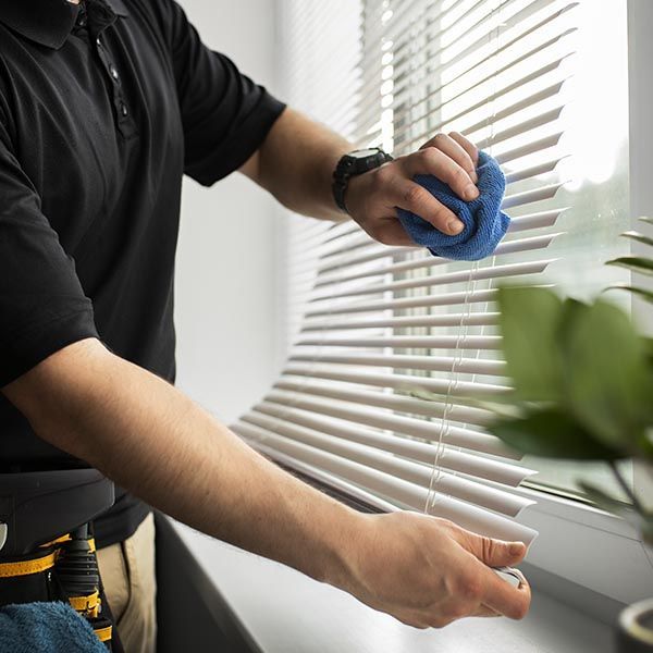 Person cleaning white blinds with a blue cloth. Window and plant visible.