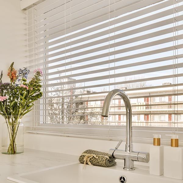 White kitchen sink and faucet with flowers, soap, and blinds over a window.