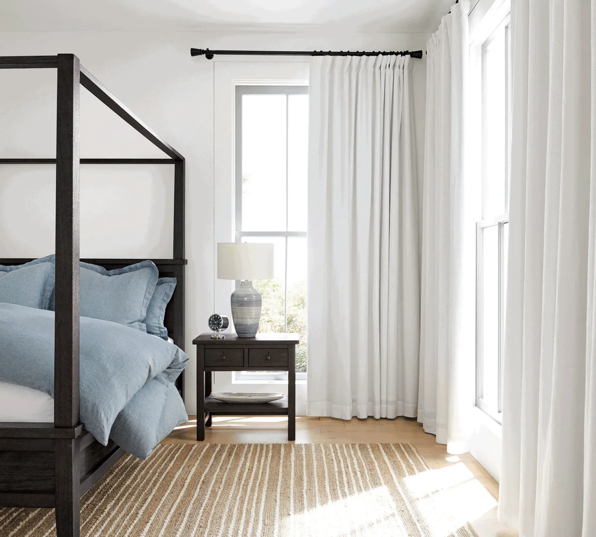 Bedroom with canopy bed, nightstand, and white curtains. Sunlight streams through the windows.