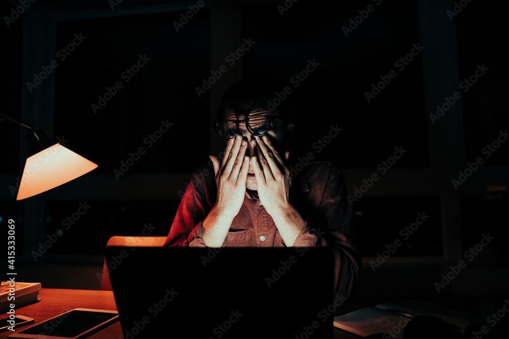 A man is sitting at a desk in front of a laptop computer covering his face with his hands.