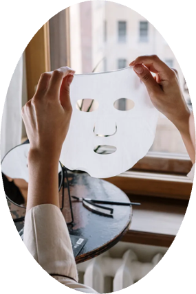 A woman is holding a face mask in front of a window.