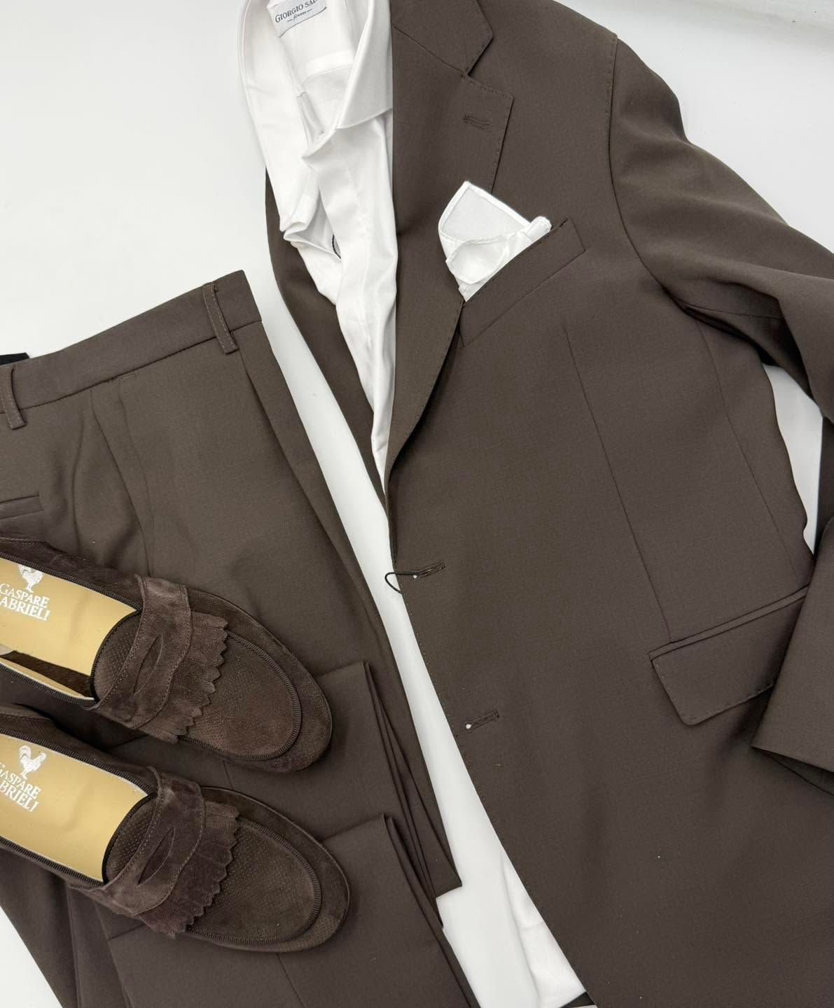 Giacca marrone, camicia bianca, fazzoletto da taschino e mocassini scuri su uno sfondo chiaro.