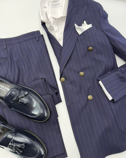 Completo blu navy a righe sottili con giacca e pantaloni, camicia bianca e mocassini neri su sfondo bianco.