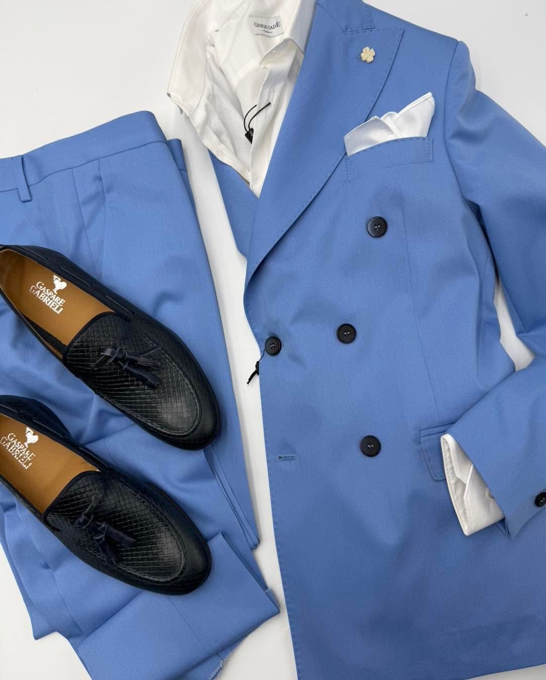 Abito blu con camicia bianca e fazzoletto da taschino, abbinato a scarpe nere su sfondo chiaro.