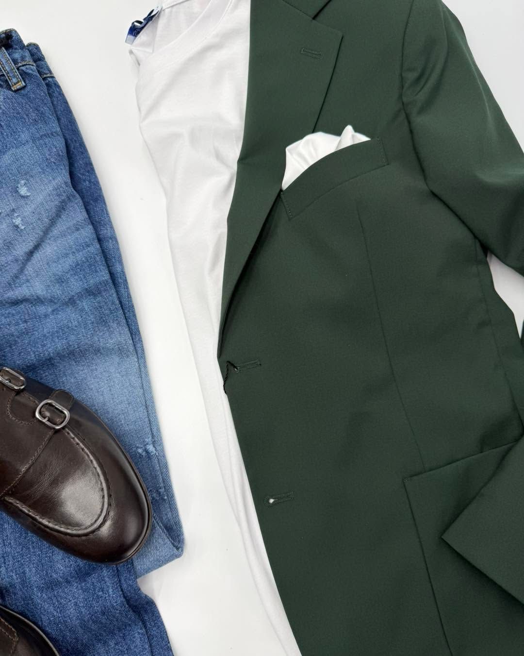 Giacca verde da completo con camicia bianca, jeans blu e mocassini in pelle marrone su sfondo piatto