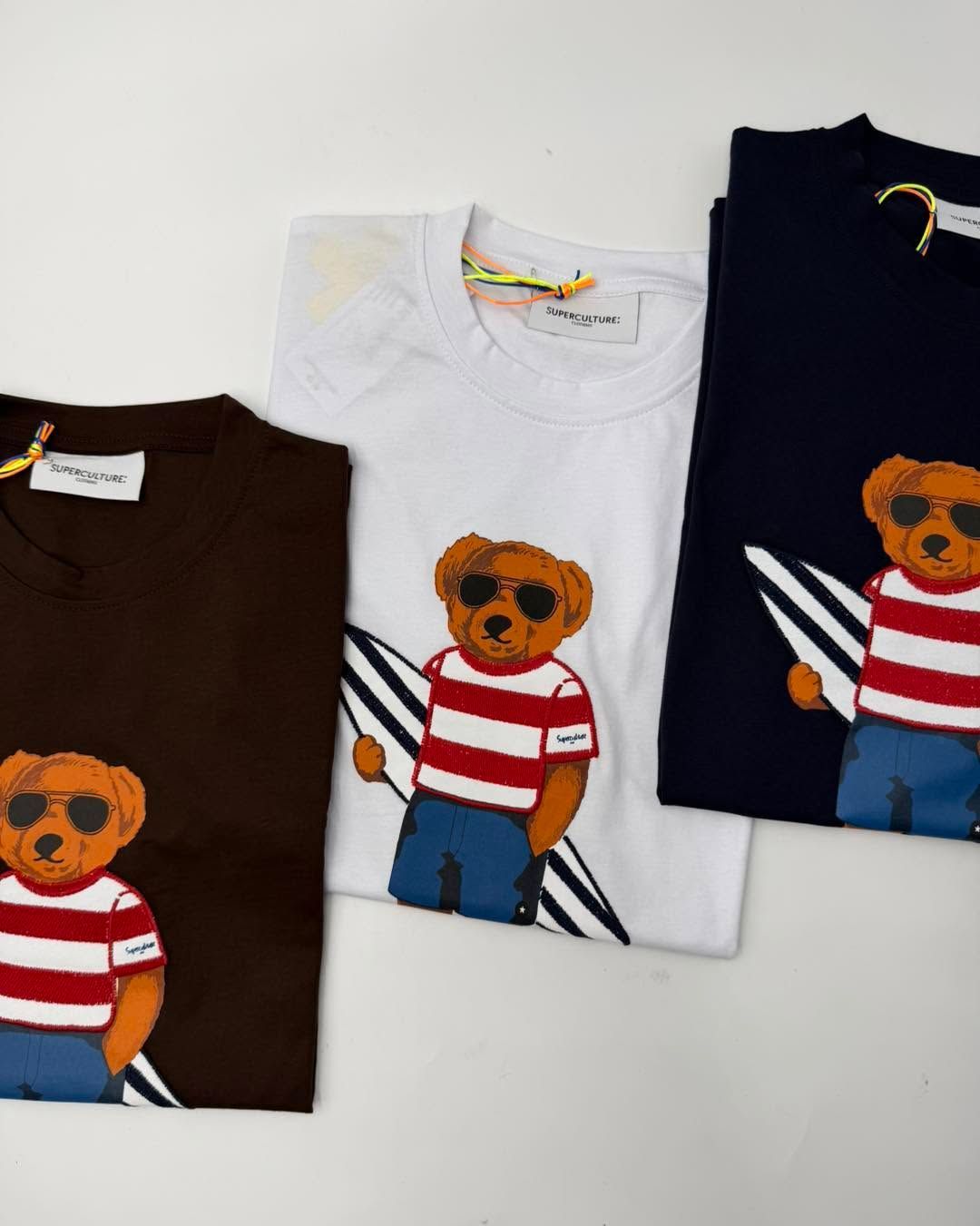 Tre magliette piegate con la stampa di un orso, nei colori marrone, bianco e blu scuro, su uno sfondo chiaro.
