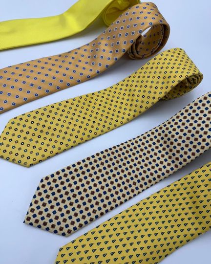 Quattro cravatte a fantasia nei colori giallo, beige e grigio, disposte in diagonale su uno sfondo chiaro.