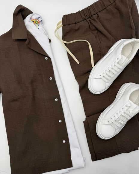 Completo composto da camicia e pantaloni marroni, steso su una superficie piana, con scarpe da ginnastica bianche.