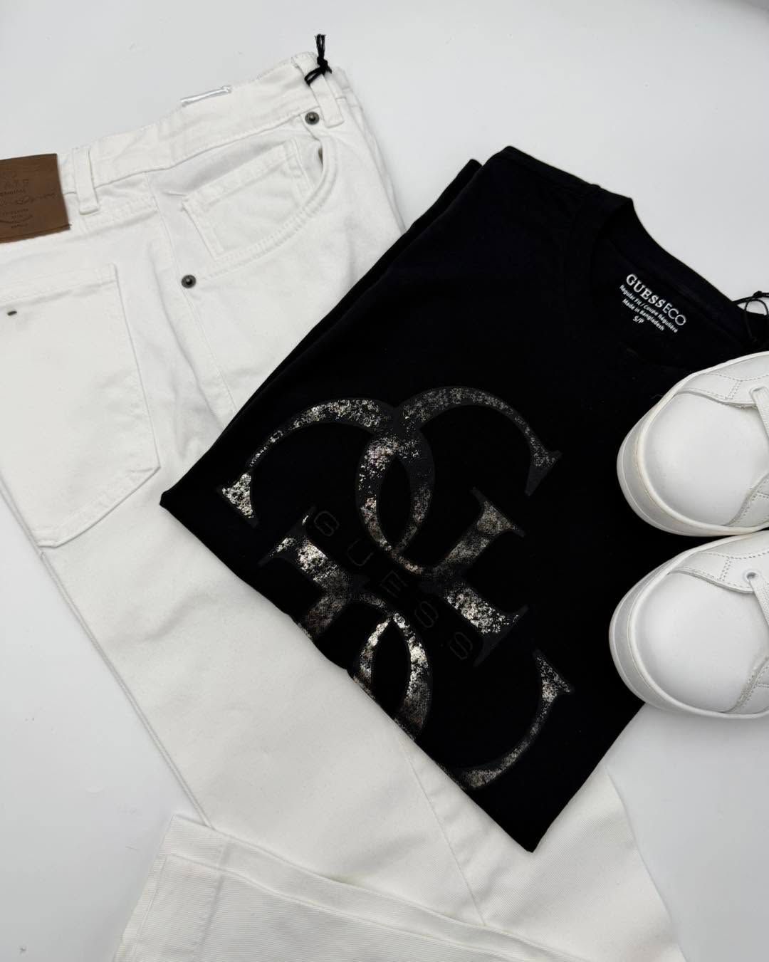 T-shirt nera con stampa grafica, jeans bianchi e scarpe da ginnastica bianche su sfondo bianco.
