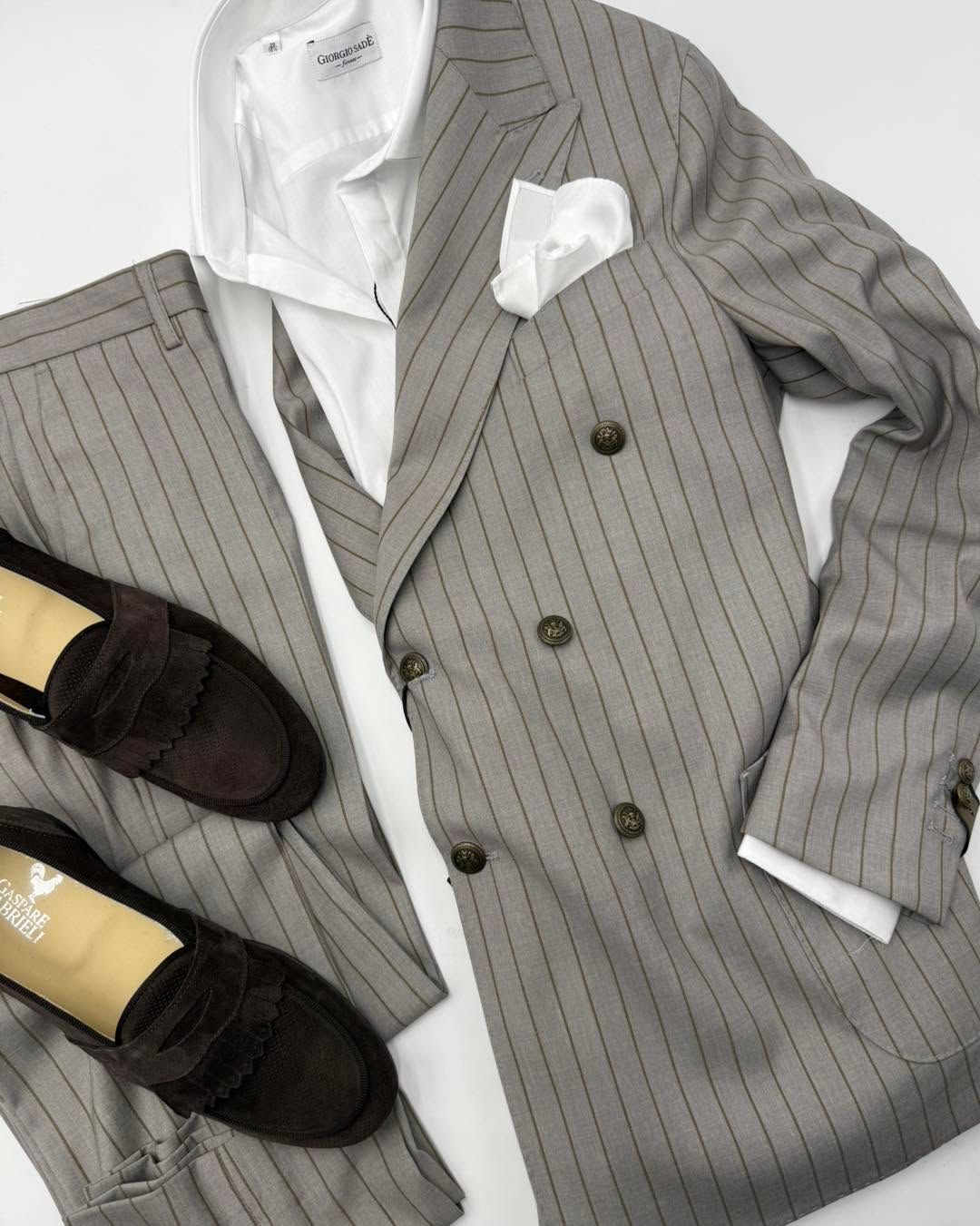 Abito gessato grigio con camicia e fazzoletto da taschino bianchi, abbinato a mocassini neri su sfondo chiaro.