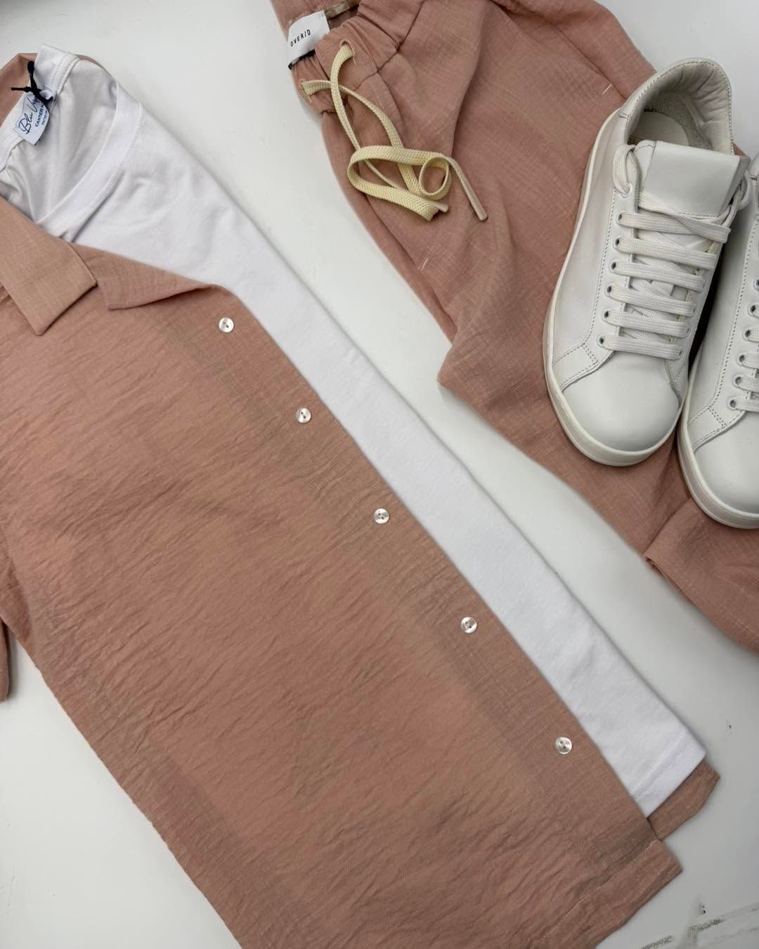 Camicia bianca e scarpe da ginnastica con un abito rosa cipria abbottonato, steso su una superficie chiara.
