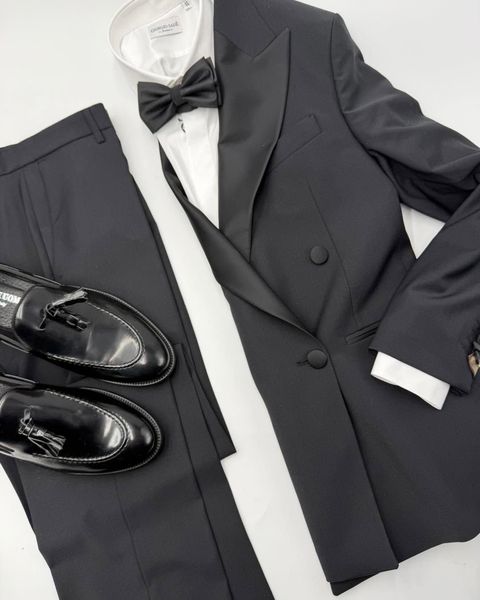 Una giacca da smoking nera, una camicia bianca, un papillon nero, pantaloni e scarpe eleganti nere lucide sono disposti su una superficie bianca.