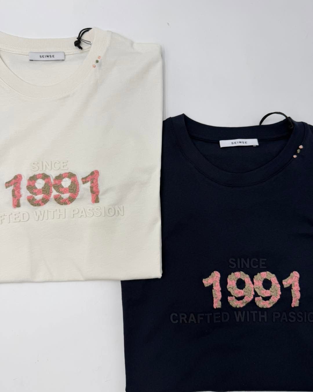 Due magliette grafiche piegate, color crema e blu navy, con la scritta rosa 