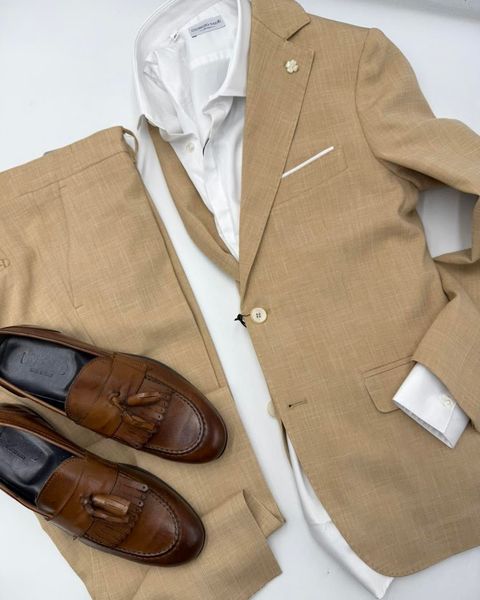 Blazer color cammello con camicia bianca, pantaloni beige e mocassini marroni, il tutto disposto ordinatamente su uno sfondo bianco.
