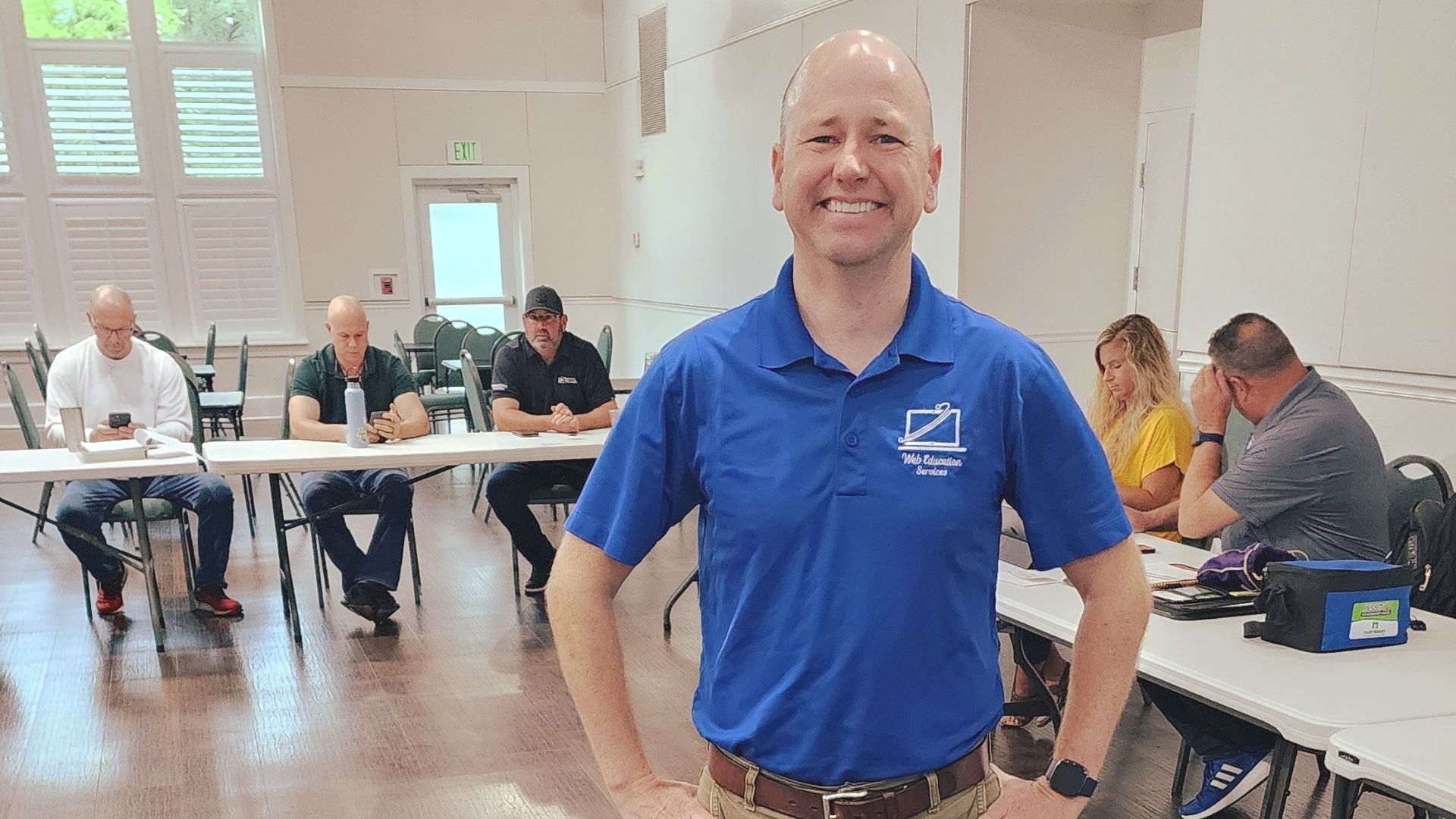 A man in a blue shirt is standing in front of a group of people sitting at tables.