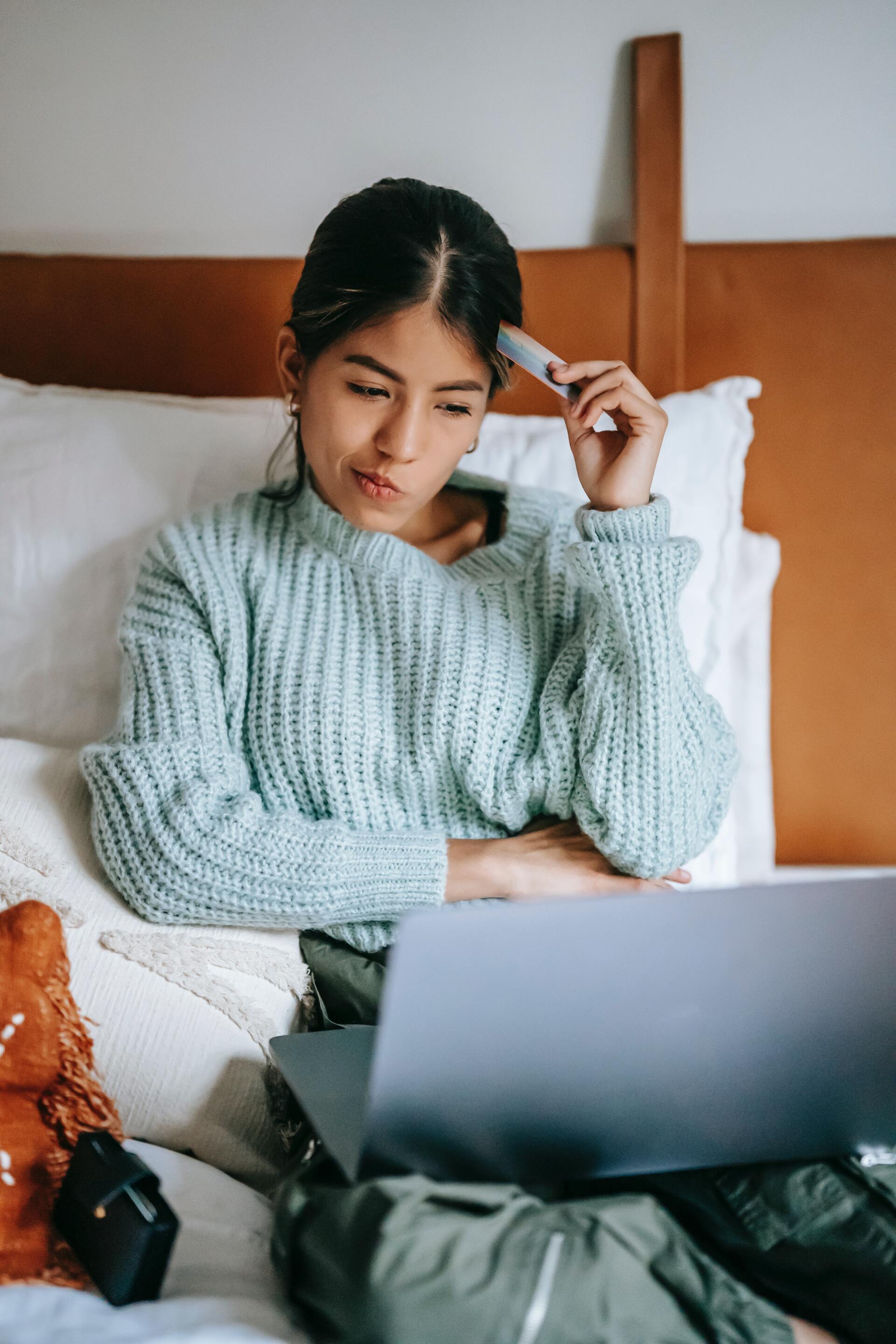 A woman is sitting on a bed using a laptop computer and holding a credit card.