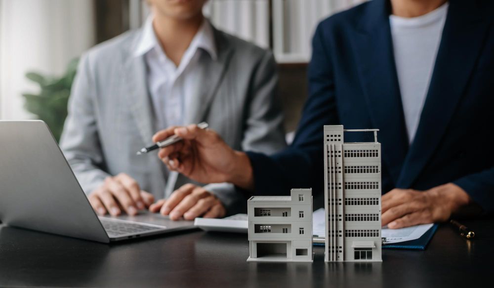 Two people are sitting at a table with a model house and a clipboard.