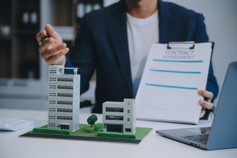 A man is sitting at a desk holding a clipboard and a model of a building.