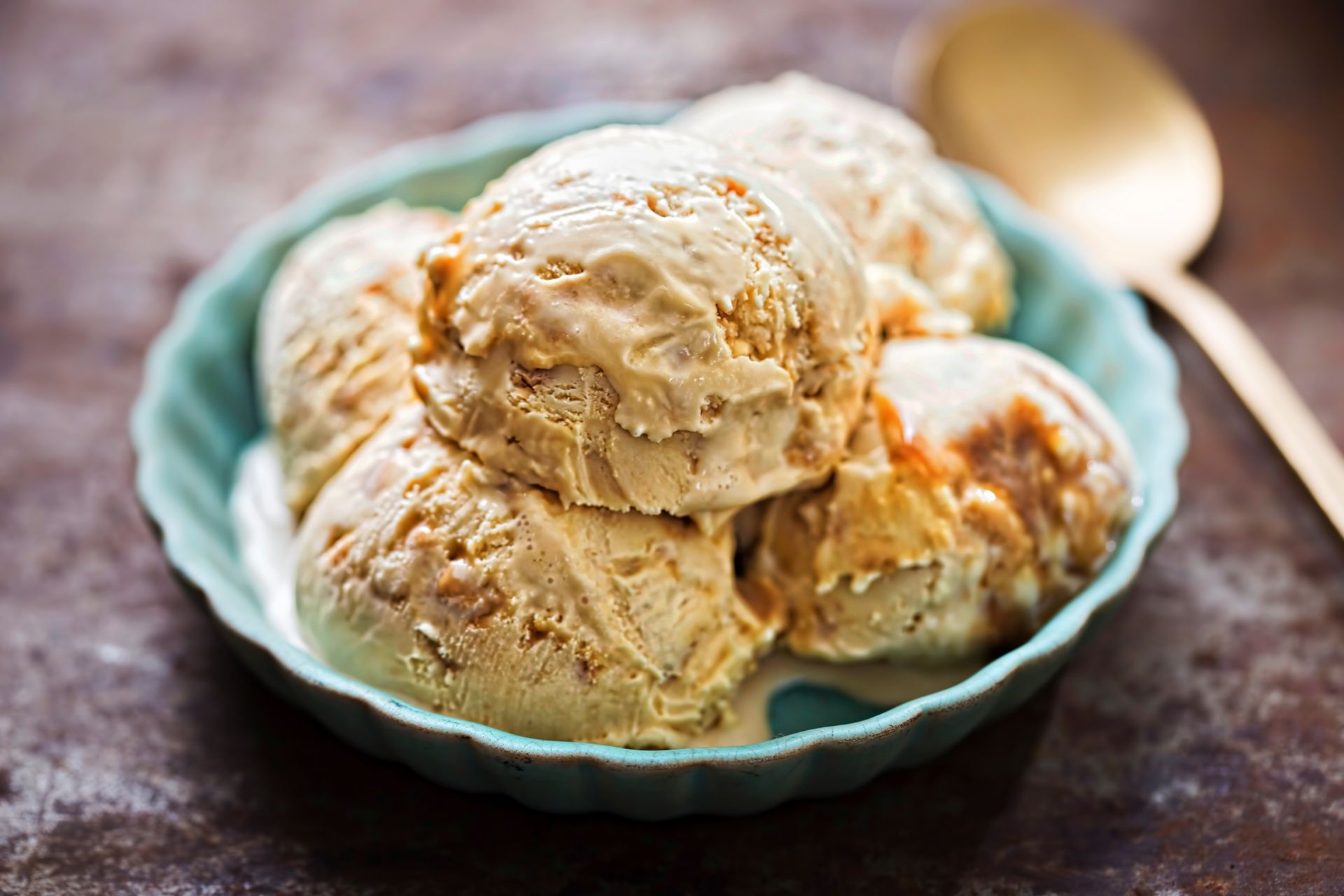 Scoops of caramel ice cream in a blue bowl, with a gold spoon on a brown surface.