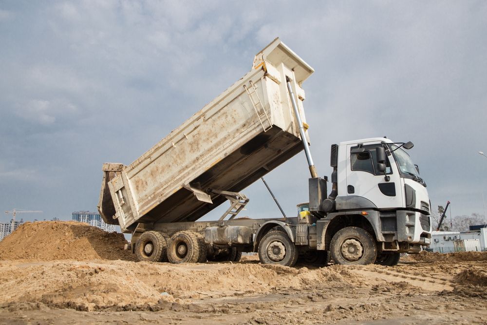 Dump truck, white cab, bed raised, unloading sand on a construction site.
