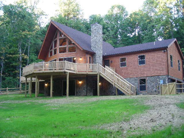 Wood-sided house with a large deck, stone chimney, and glass windows set in a grassy yard surrounded by trees.