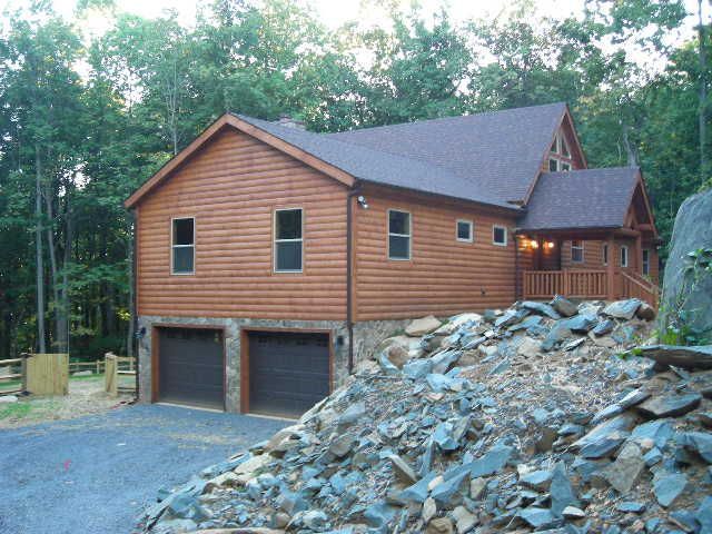 Brown wood cabin with two garage doors, set in a wooded area with a stone pile in the foreground.