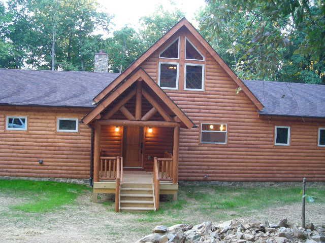 Log cabin with wooden porch and triangular window. Brown siding, set in a grassy yard, surrounded by trees.