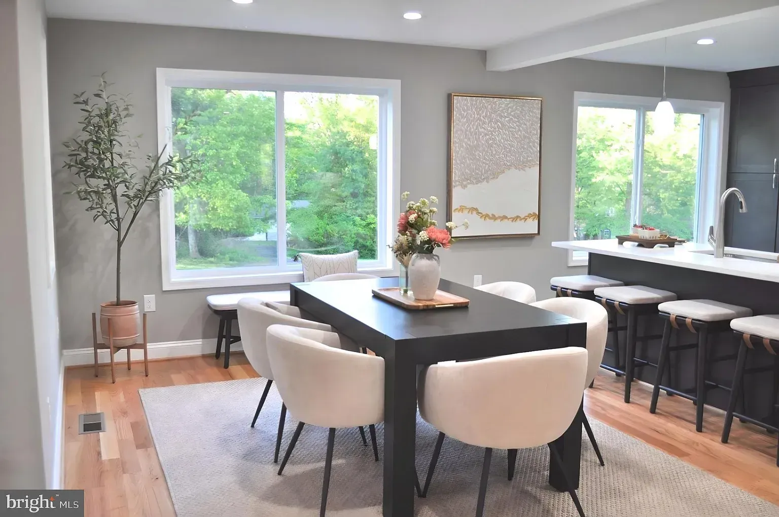 Dining room with a dark table, white chairs, and large windows overlooking trees.