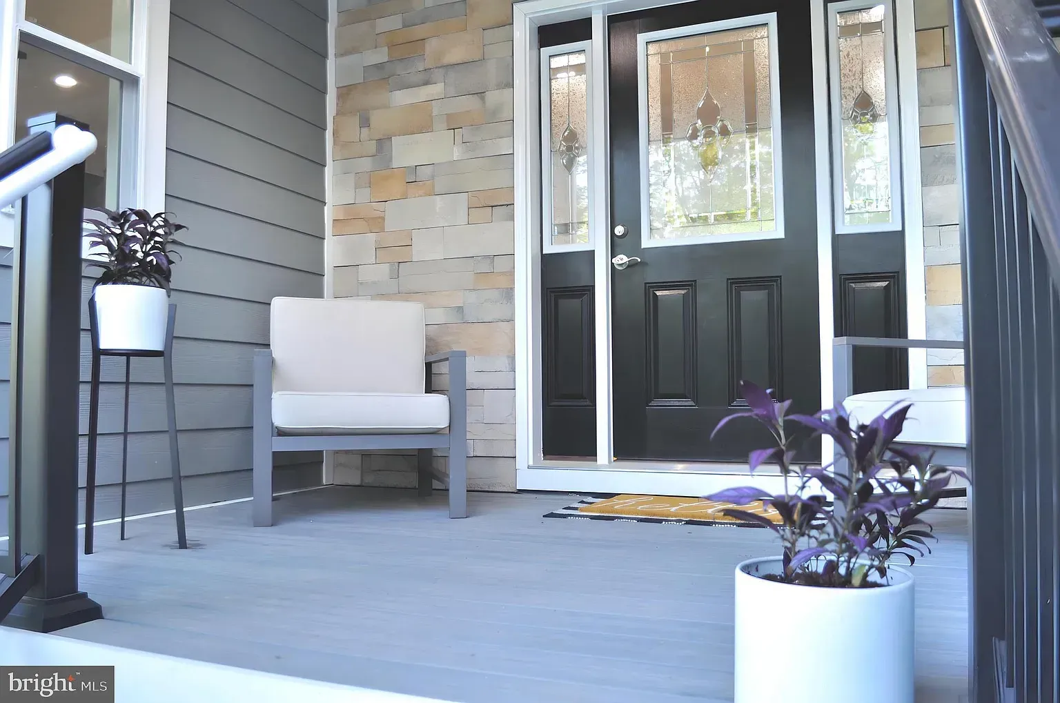 Front porch with gray siding, stone accent wall, black door, chair, potted plants.