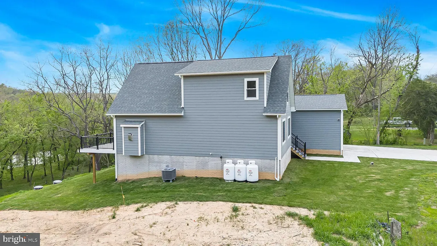 Gray house with attached shed and propane tanks on a grassy hill under a blue sky.