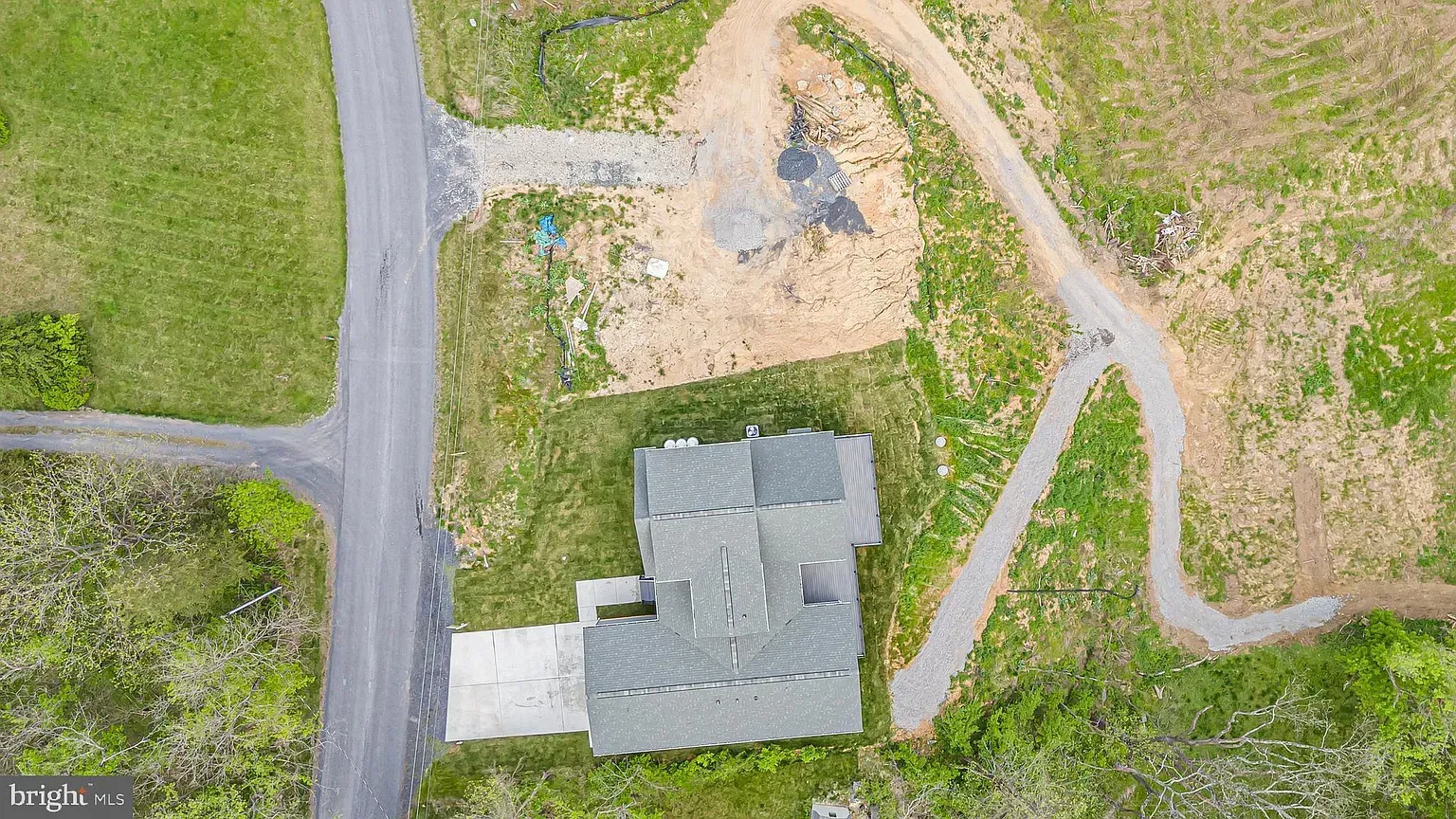 Aerial view of a house with gray roof, concrete driveway, and surrounding green grass and dirt roads.