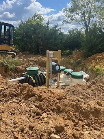 Green septic tanks in an excavated area. Dirt surrounds the tanks and plumbing. Trees and field in the background.