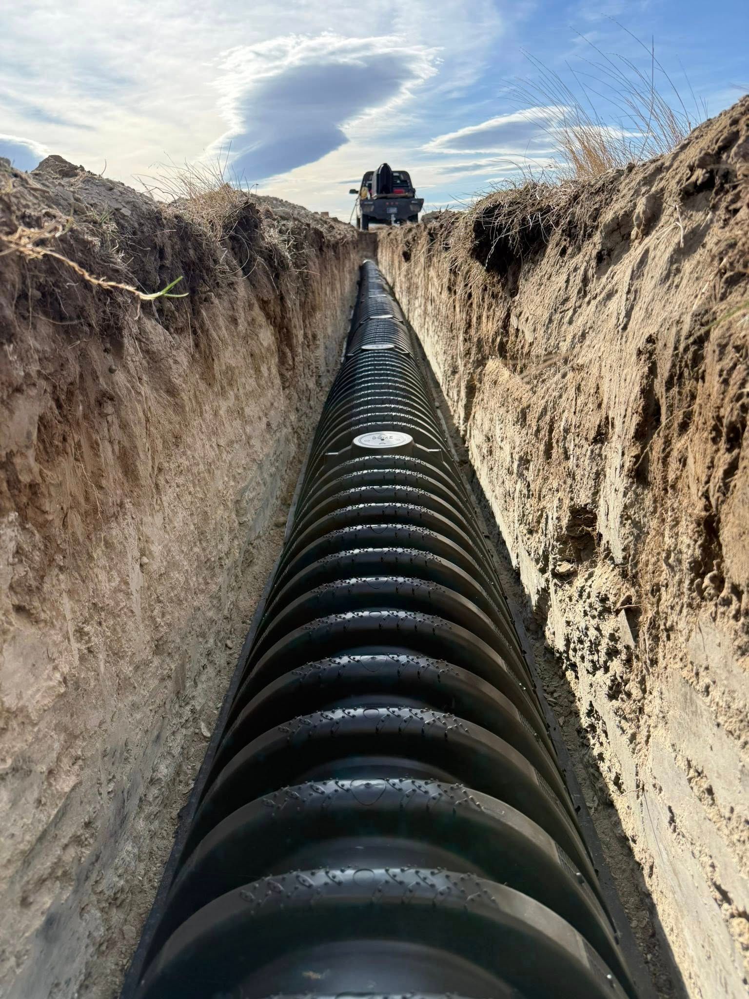 Large black pipe running through a dirt trench toward a machine under a cloudy sky