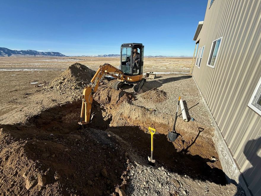 Excavator digging near a house foundation in a dirt trench under a clear sky
