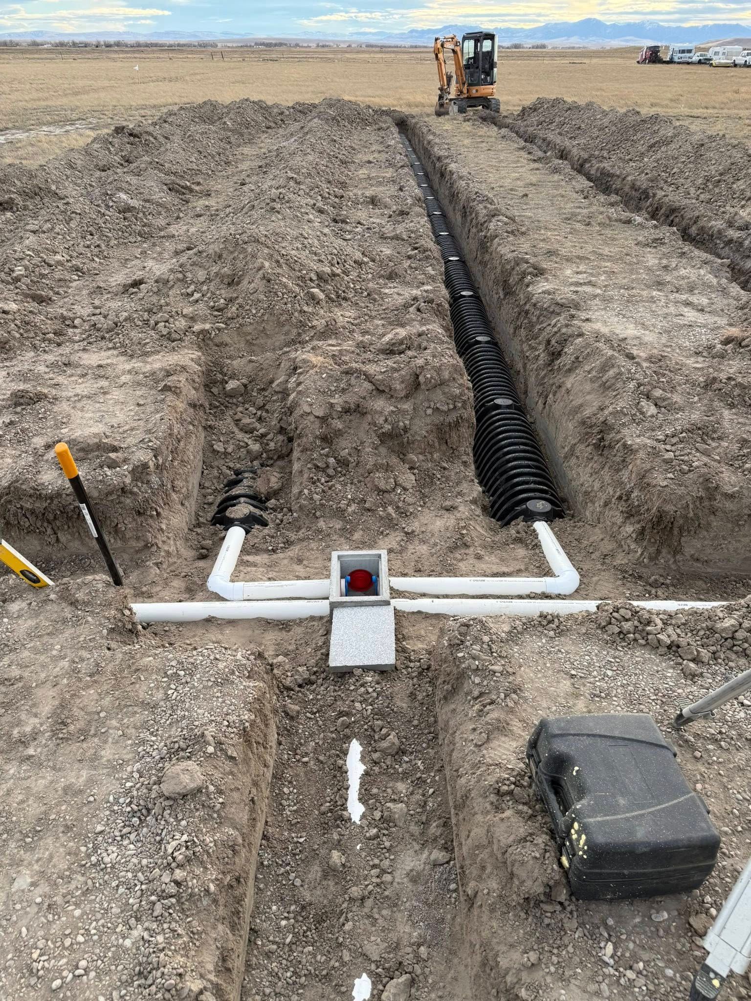 Trench with buried utility conduit and junction box at a construction site, with a loader in the distance