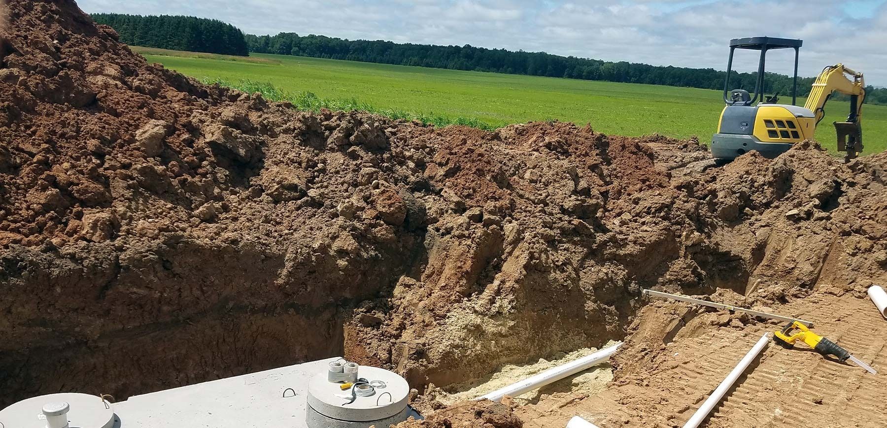 Construction excavation pit with dirt piles, exposed pipes, and an excavator in a grassy field