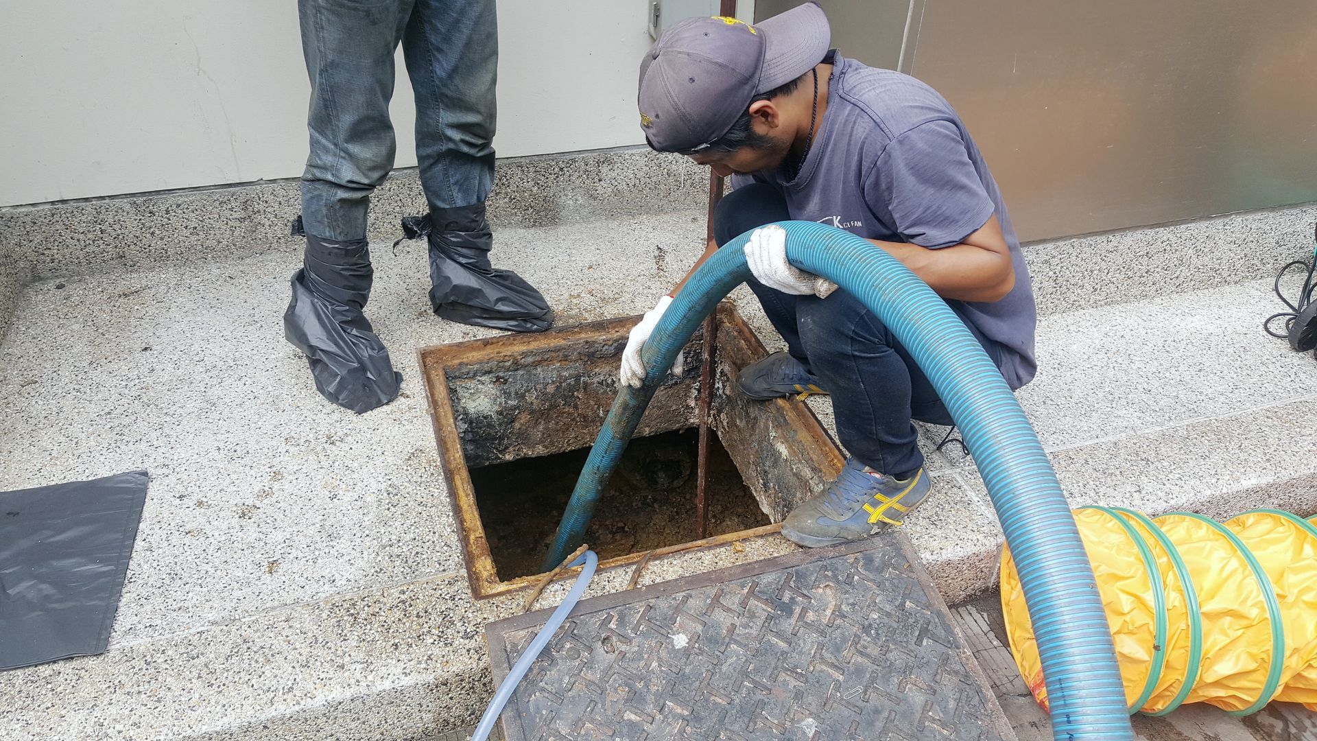 Worker cleaning a drainage opening with a hose beside a building wall