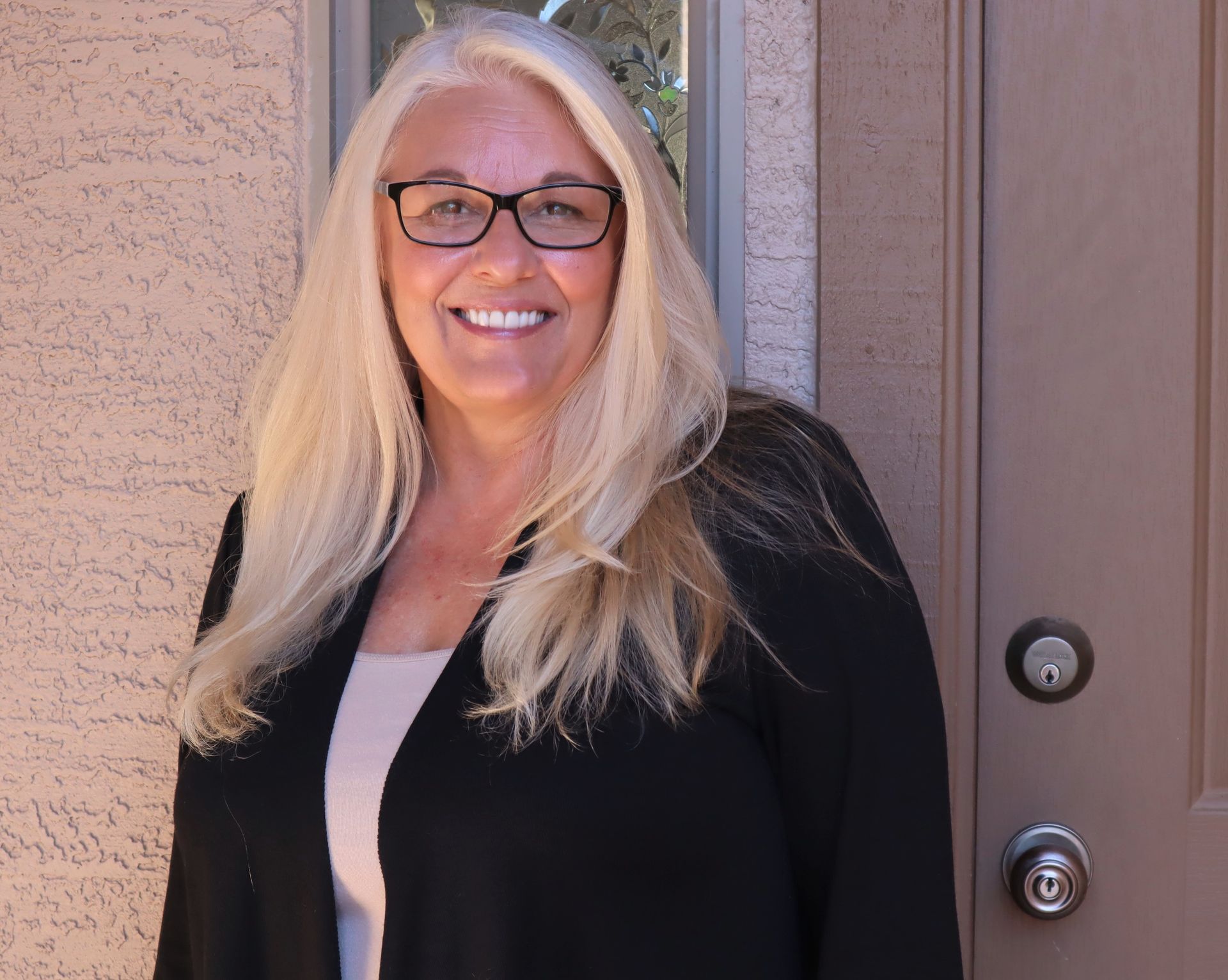 Woman with glasses smiles in front of a tan wall and brown door. She wears a black cardigan over a light shirt.