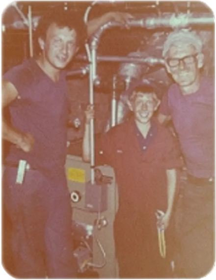 Three people in work uniforms stand near equipment. Two smile, and one holds a tool. The setting is indoors.