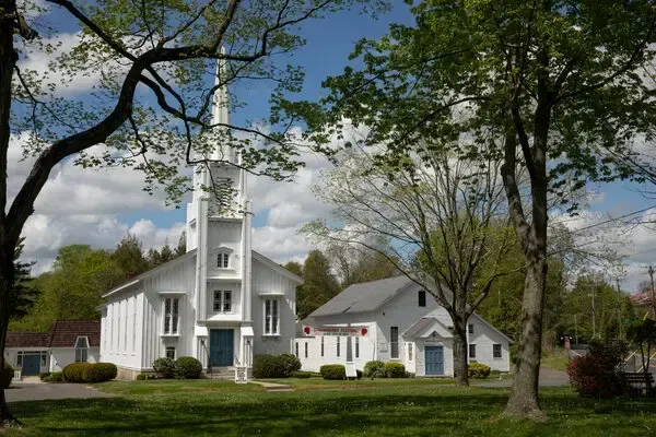 White church with tall steeple and small white building, framed by trees under a blue cloudy sky.