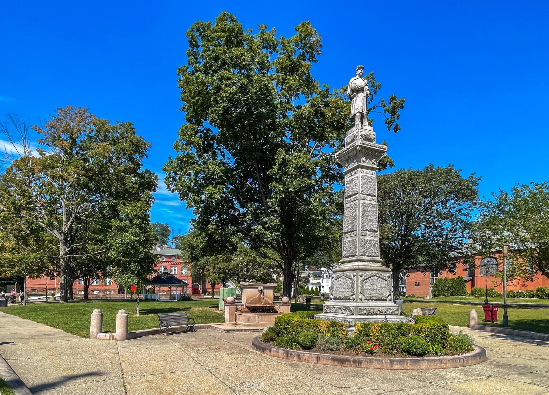 Stone monument statue in a sunny park with trees, grass, and a bright blue sky