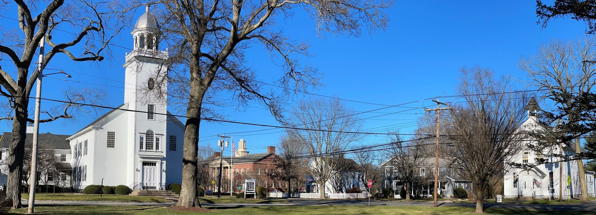 White church with a tall steeple beside a tree-lined street under a clear blue sky.