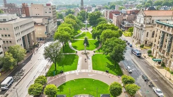 Aerial view of a green city park with paths, trees, and surrounding buildings and streets.