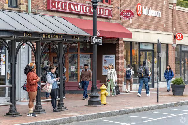 People standing outside storefronts on a city sidewalk, with a red awning and yellow fire hydrant.