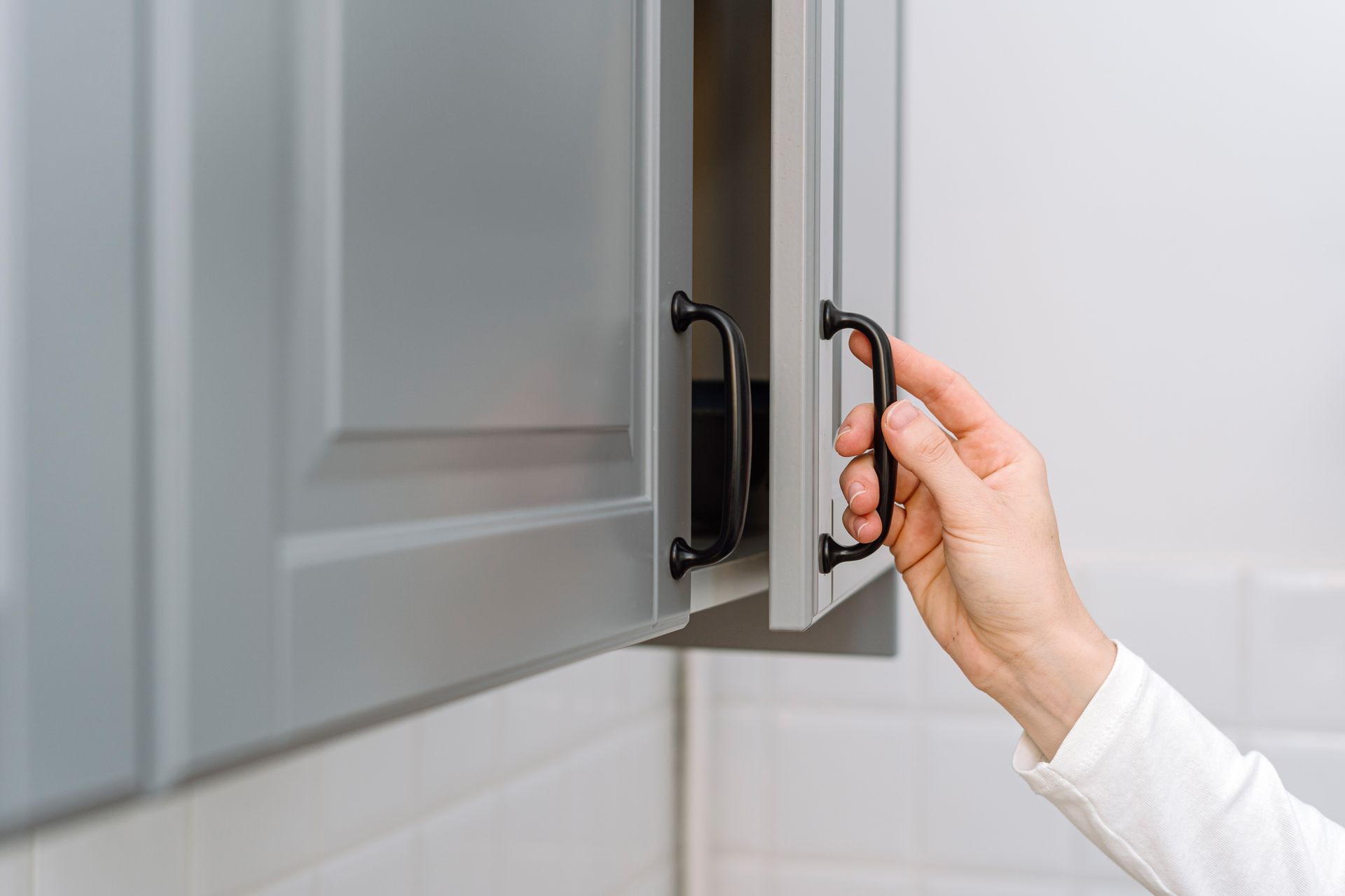 Hand opening a gray cabinet door by its black handle in a bright kitchen.