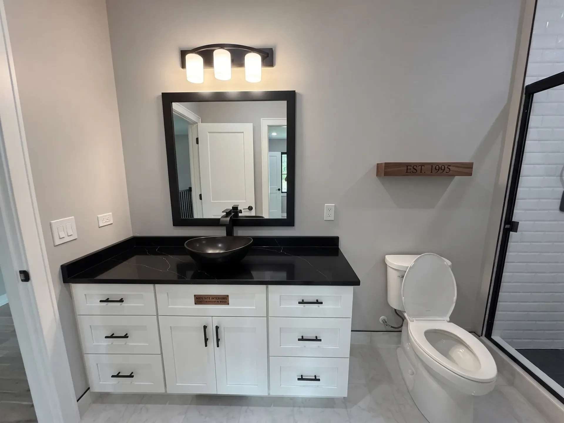 Modern bathroom with white vanity, black countertop, mirror, wall light, and toilet beside a glass shower.