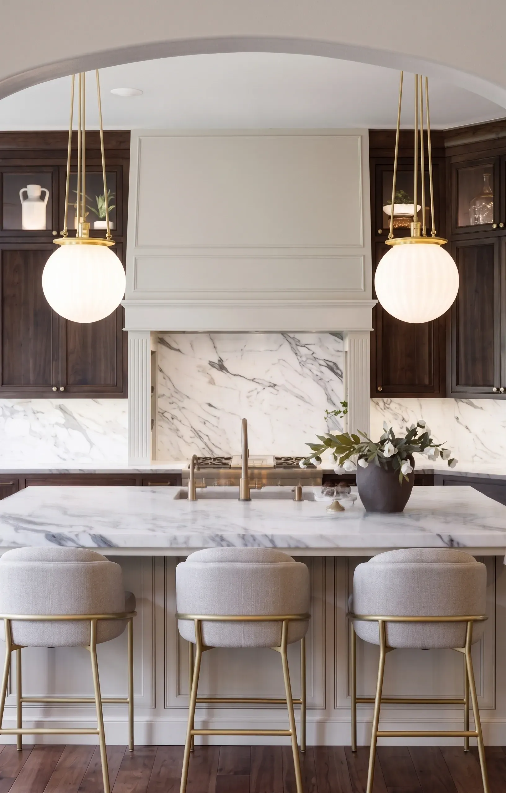Modern kitchen with marble island, gold pendant lights, dark cabinets, and upholstered stools.