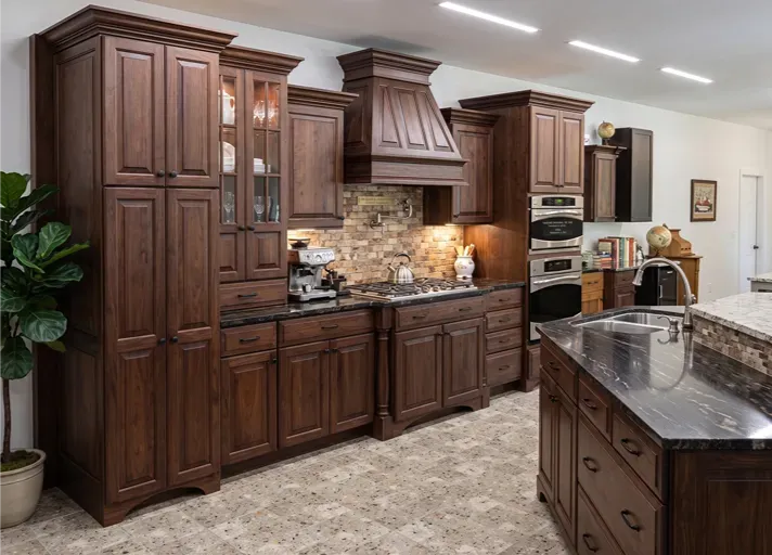 Dark wood kitchen with granite island, stainless appliances, and stone backsplash under bright ceiling lights.
