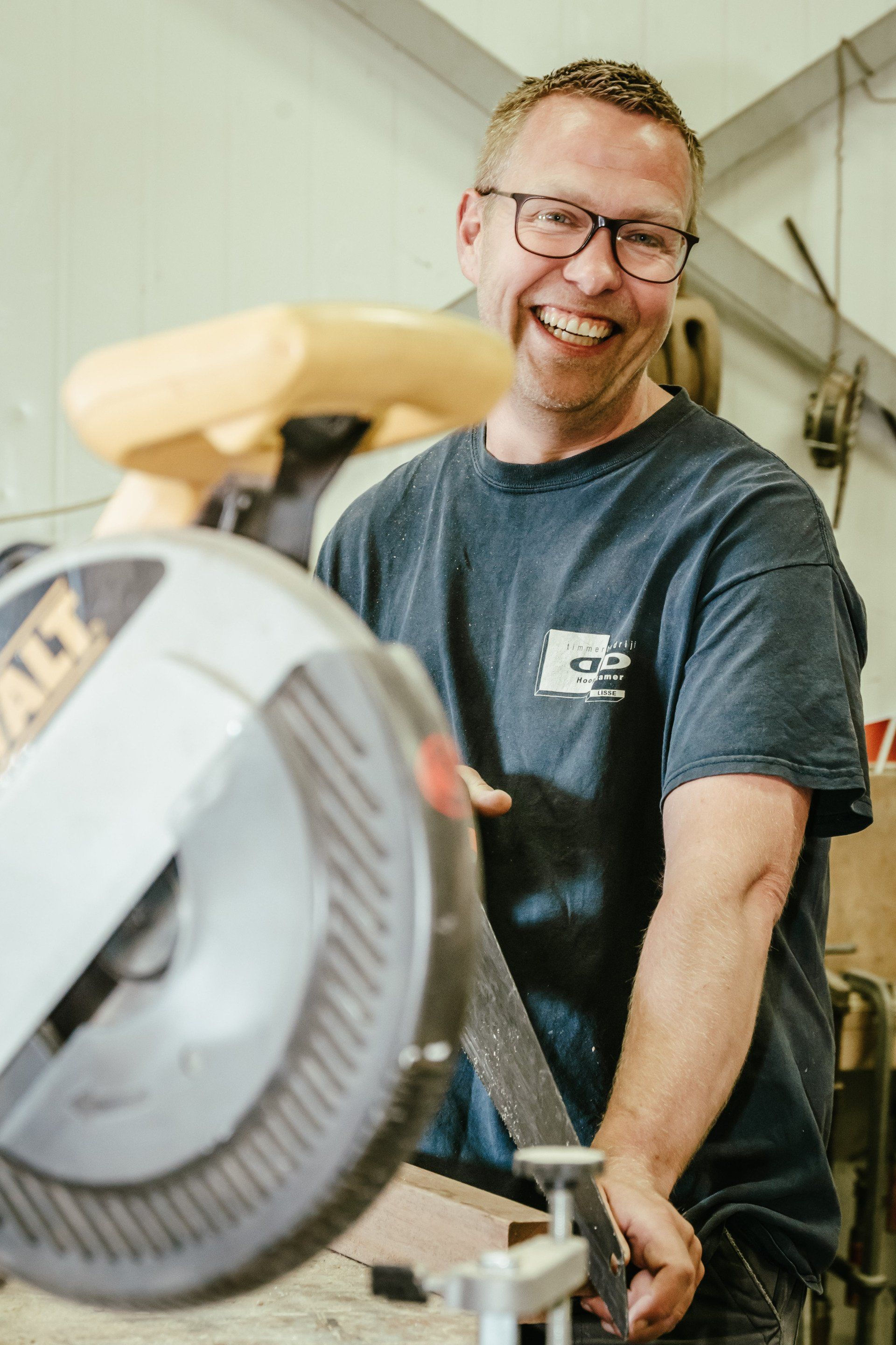 Lachende man met bril zaagt hout met een elektrische zaag in een werkplaats.